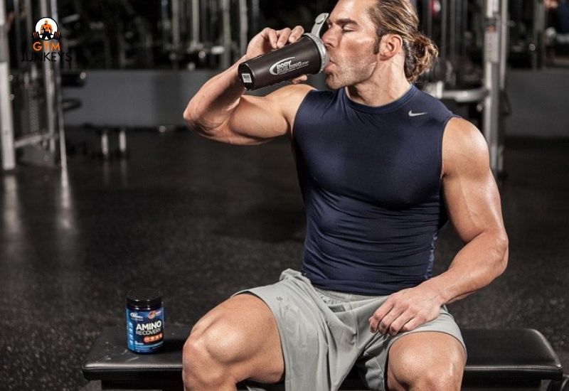 A muscular man drinks amino acid after a workout while sitting on a gym bench.